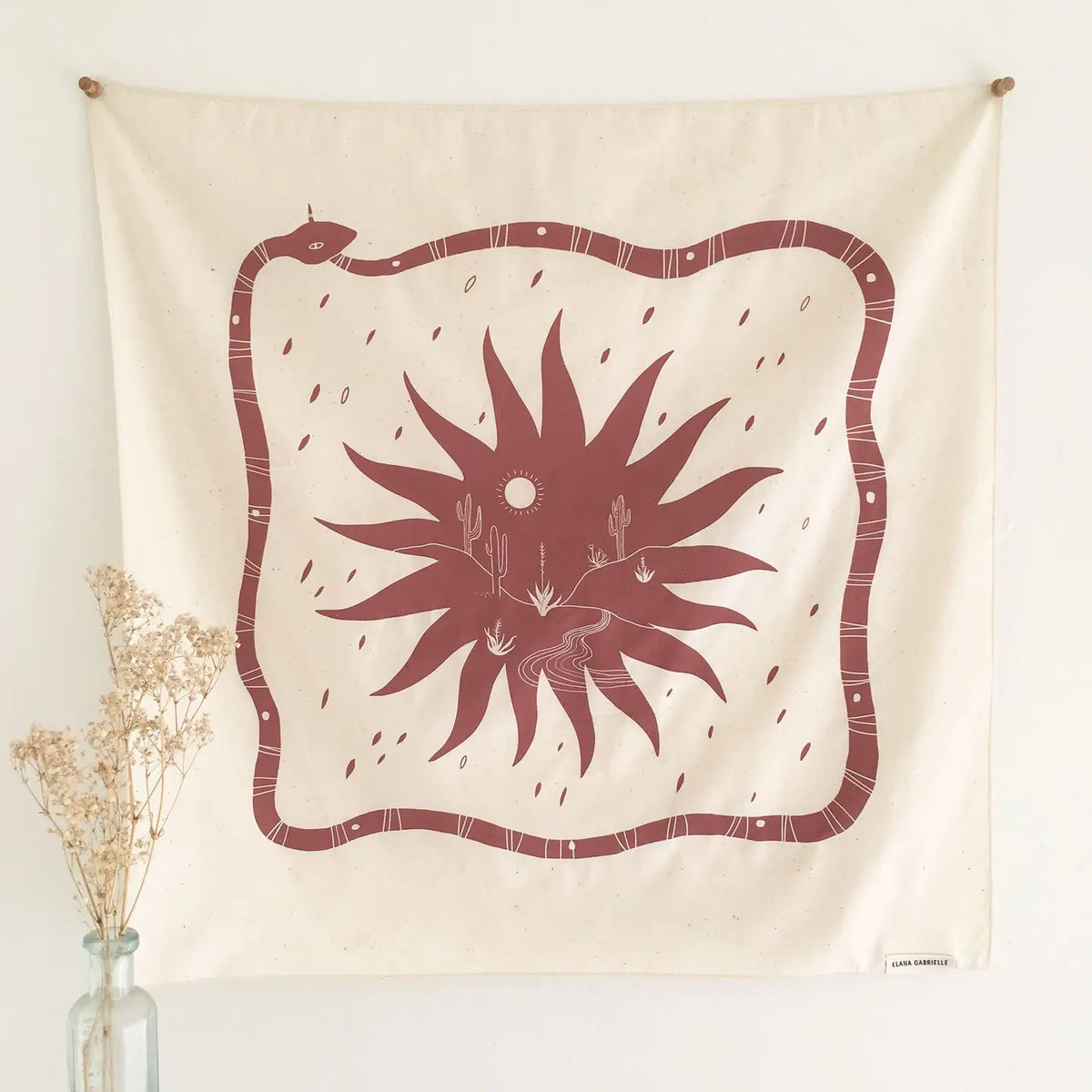 Cream Elana Gabrielle Desert Bandana pinned flat to a white wall, showing its terracotta snake border and central sunburst desert scene, with a clear vase of dried white flowers at lower left.