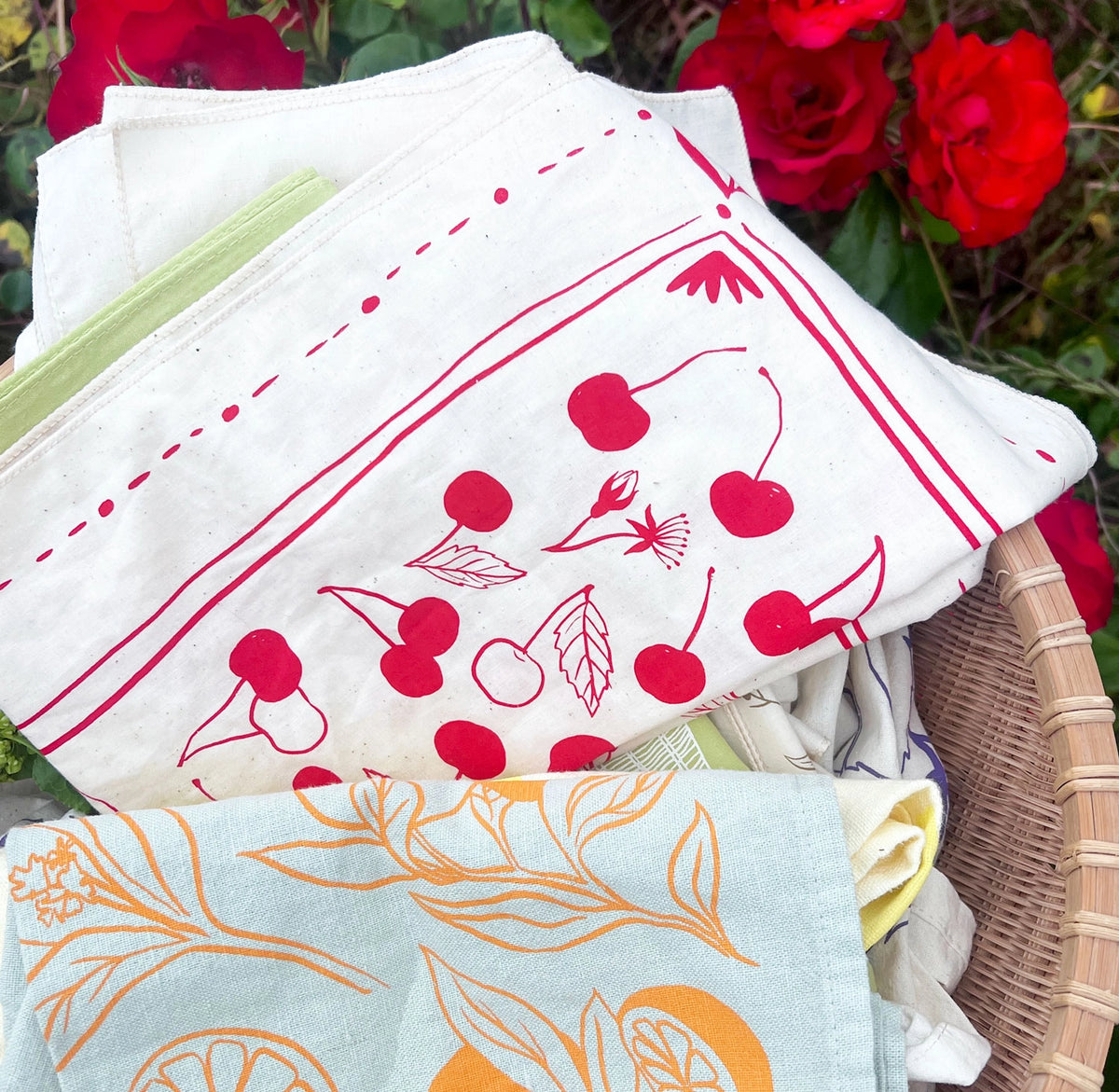 Cream Elana Gabrielle Cherries Bandana folded in a woven basket among other printed textiles, with bright red cherry motifs in front of blooming red roses.