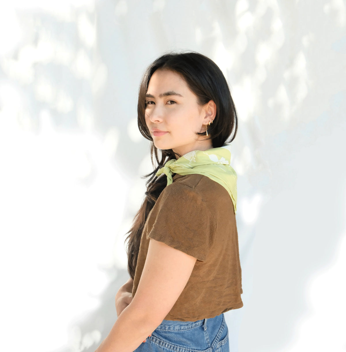 Woman in a brown T-shirt and jeans wearing the chartreuse Elana Gabrielle Forage Bandana tied around her neck, with white botanical print visible in soft dappled light.