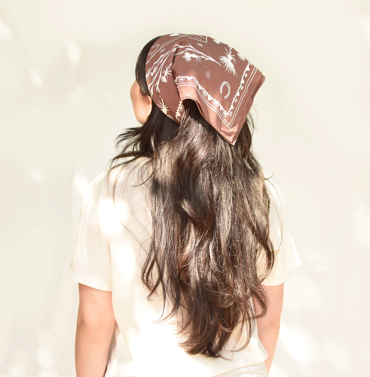 Woman seen from behind wearing the chocolate-brown Elana Gabrielle Pastures Bandana tied over her long dark hair as a headscarf, with white horseshoe and thistle motifs visible.