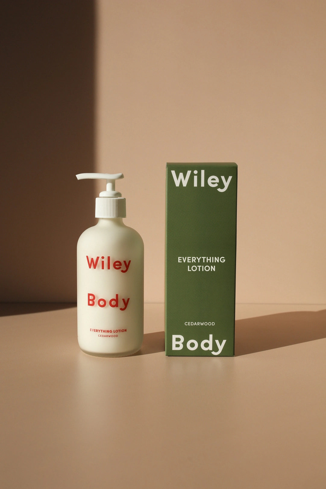 Wiley Body Everything Lotion Cedarwood in an off-white pump bottle with red lettering, standing beside a tall olive-green carton with white branding in warm directional sunlight.