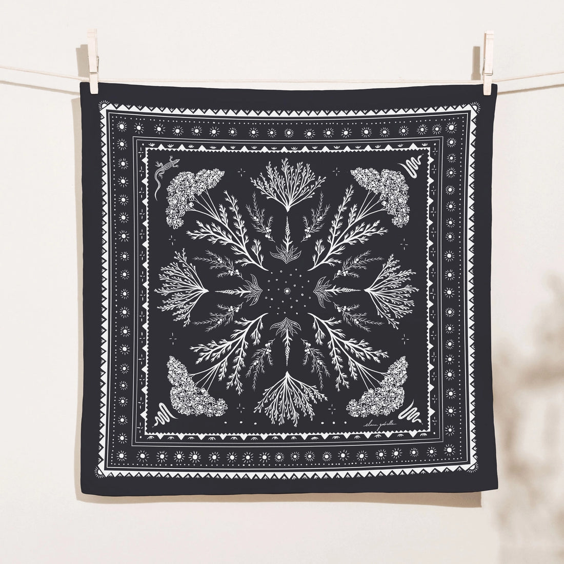 Black square High Desert Bandana clipped to a clothesline, printed in white with a symmetrical botanical design of desert plants, flowering clusters, snakes, suns and decorative geometric borders.