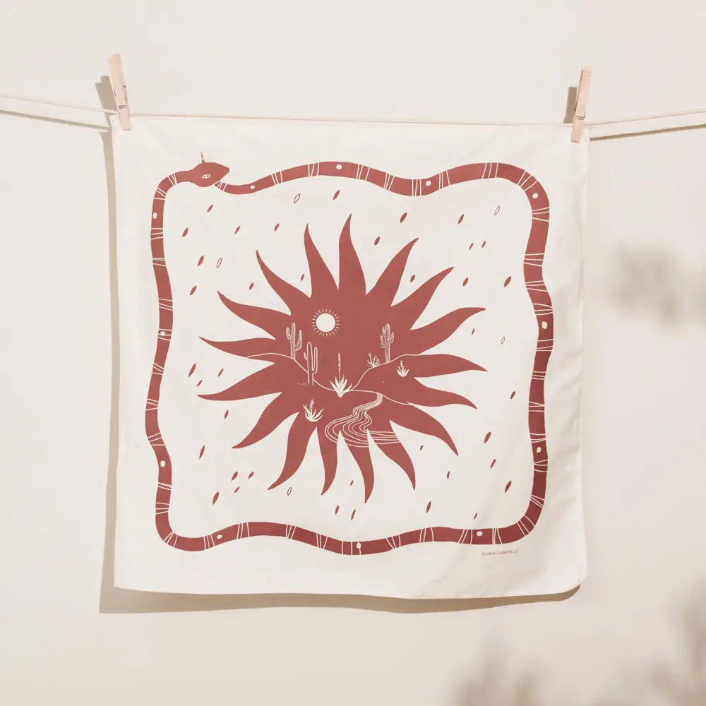 Cream Elana Gabrielle Desert Bandana clipped to a clothesline, screen printed in terracotta with a long snake border surrounding a large sunburst and tiny desert landscape of cacti and a winding path.