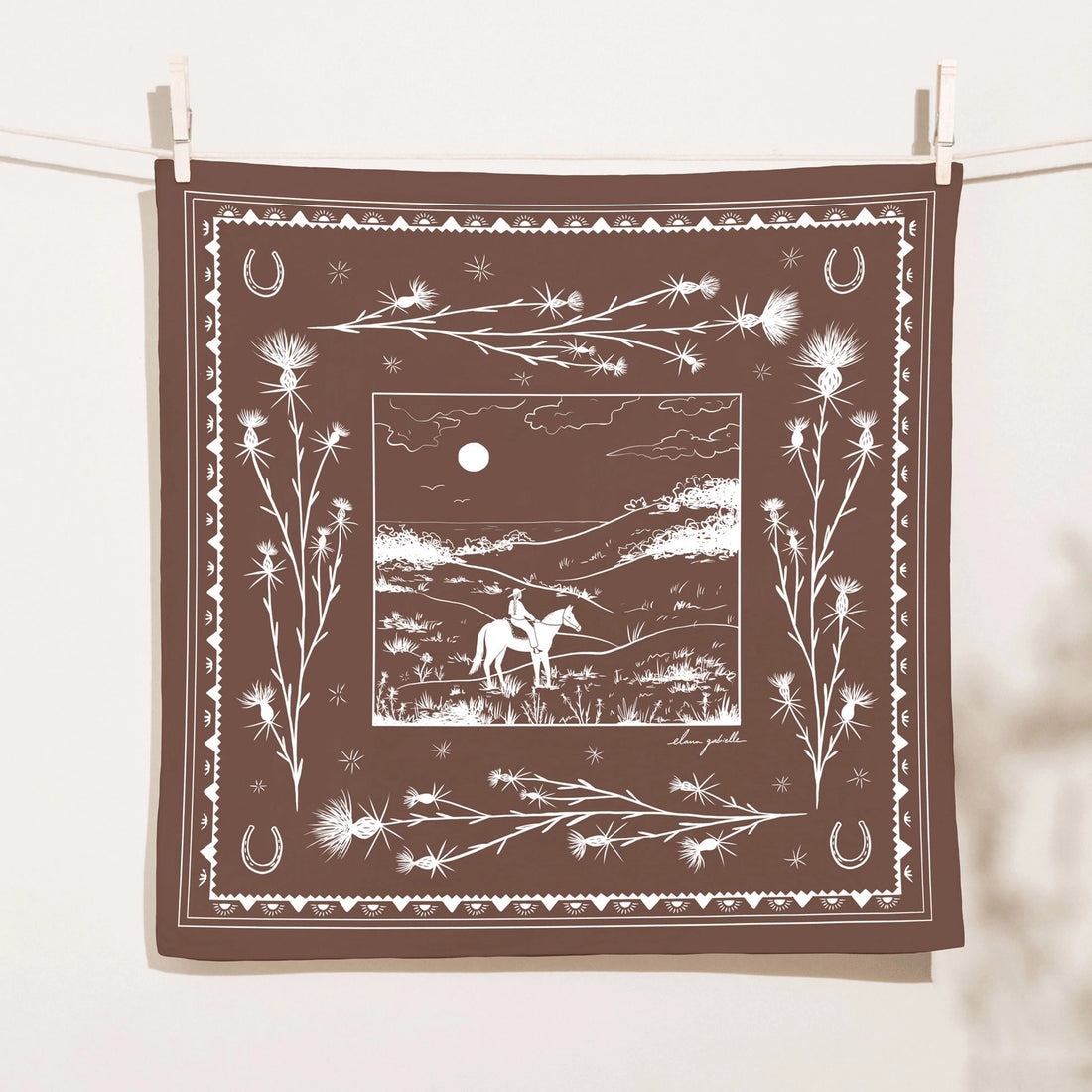 Chocolate-brown Elana Gabrielle Pastures Bandana clipped to a clothesline, screen printed in white with a moonlit horseback rider, thistles, horseshoes and decorative Western border motifs.
