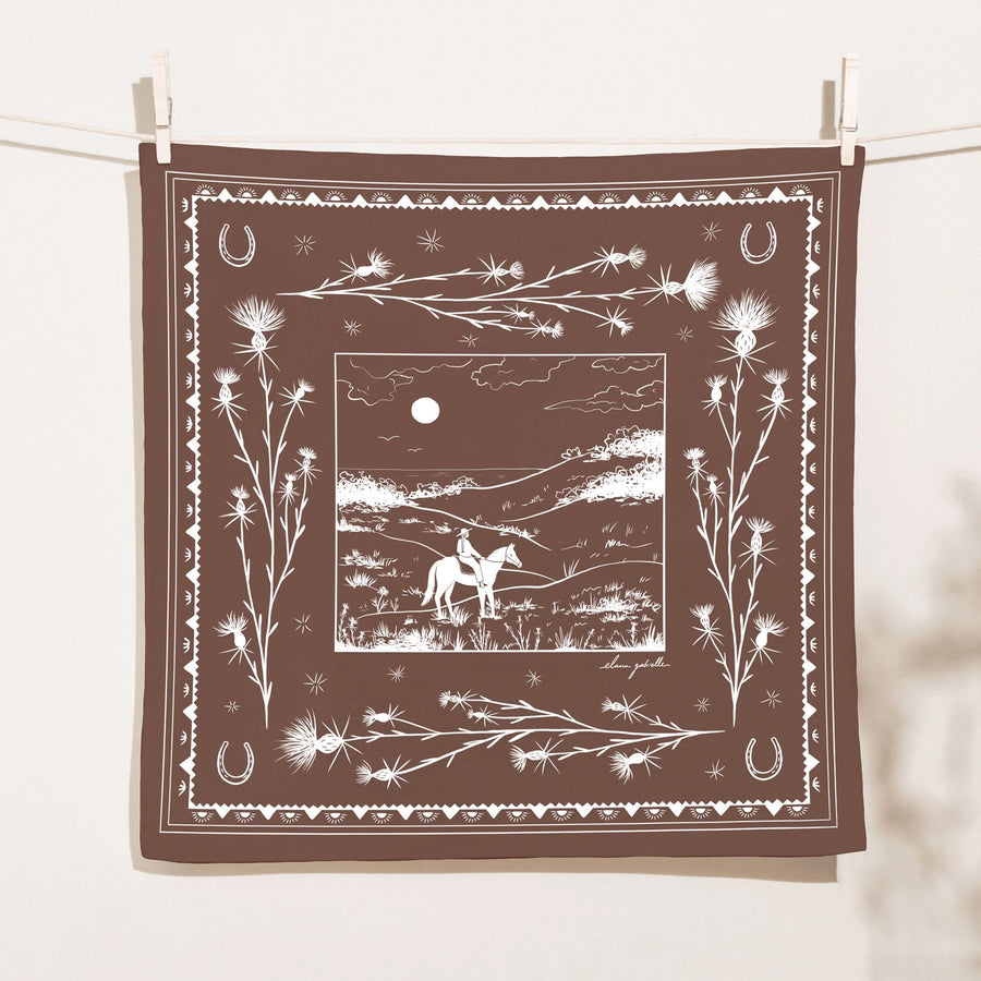 Chocolate-brown Elana Gabrielle Pastures Bandana clipped to a clothesline, screen printed in white with a moonlit horseback rider, thistles, horseshoes and decorative Western border motifs.