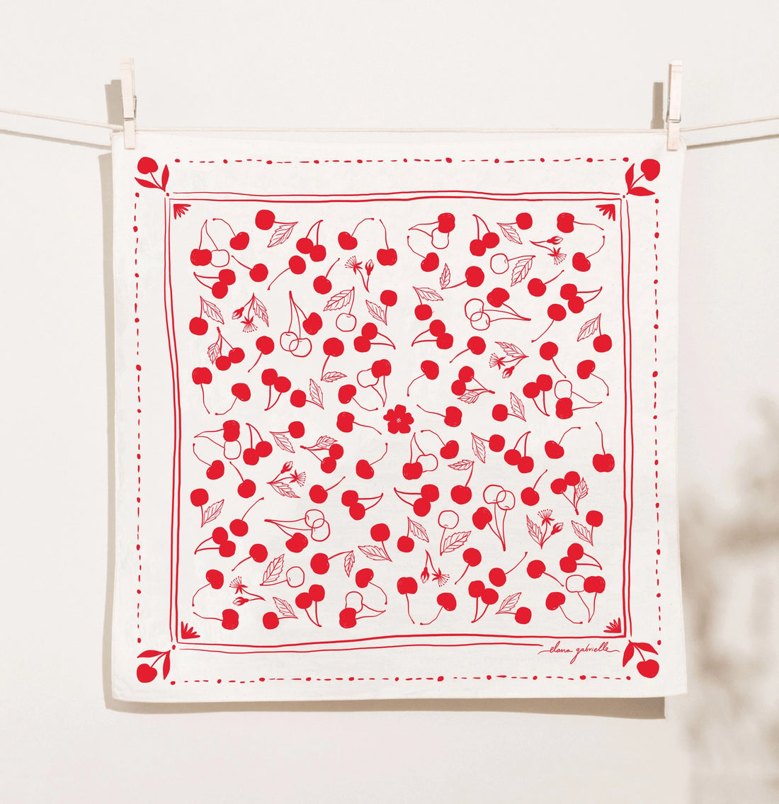 Cream Elana Gabrielle Cherries Bandana clipped to a clothesline, screen printed in bright red with scattered cherries, leaves and small blossoms inside a hand-drawn cherry border.