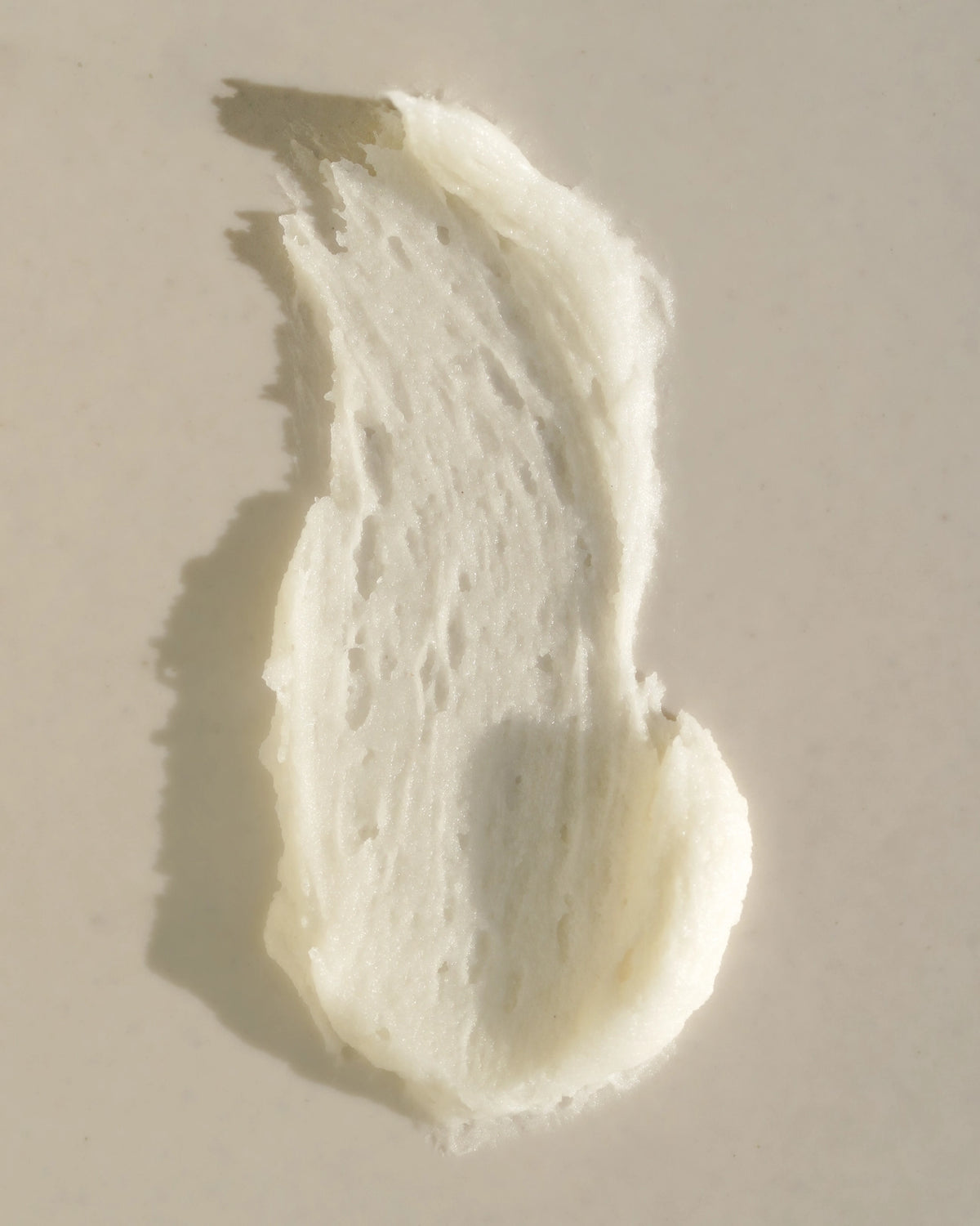 Close-up smear of white whipped deodorant spread across a warm beige surface, showing its dense creamy texture and small air pockets in directional sunlight.
