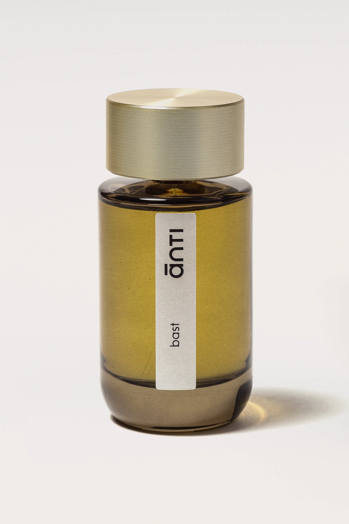 ānti Bast eau de parfum in a smoky olive glass bottle with a long vertical cream label and wide champagne-gold cap embossed with concentric ripple lines on an off-white background.