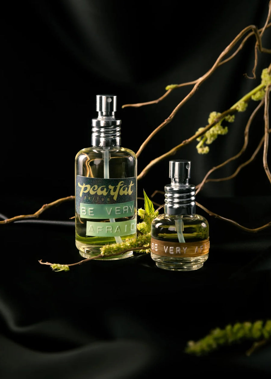 Two sizes of Pearfat Be Very Afraid perfume with exposed silver sprayers and pale green liquid, arranged among twisting bare branches and tiny green flowers against draped black fabric.
