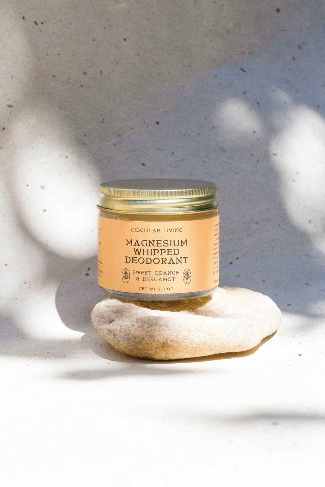 Circular Living Magnesium Whipped Deodorant, Sweet Orange & Bergamot in a small clear glass jar with orange label and gold screw lid, balanced on a pale stone in dappled sunlight.