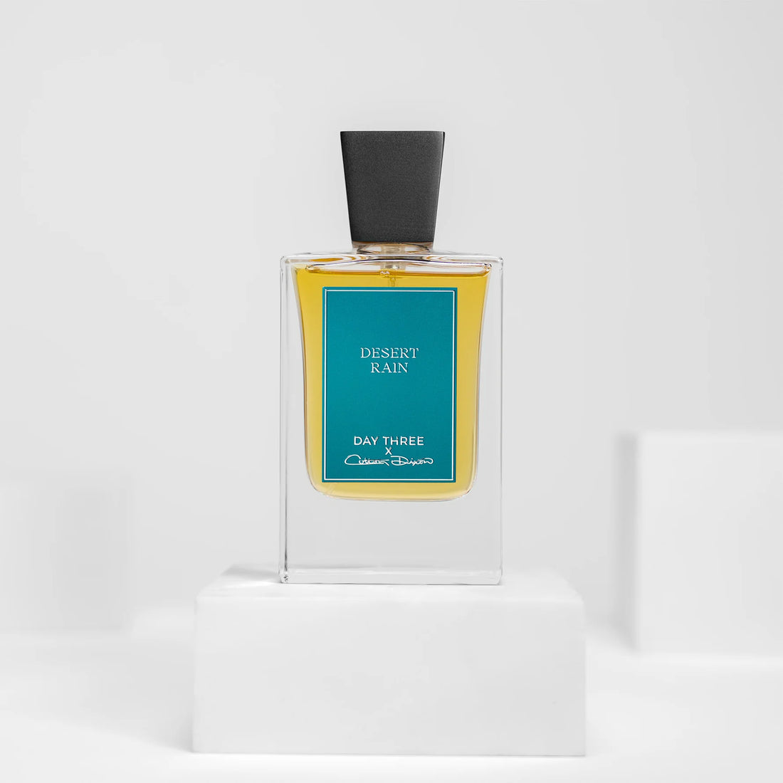 Day Three Desert Rain eau de parfum in a tall clear rectangular bottle with golden liquid, a wide angular matte-black cap and teal-blue rectangular label, centered on a white pedestal.