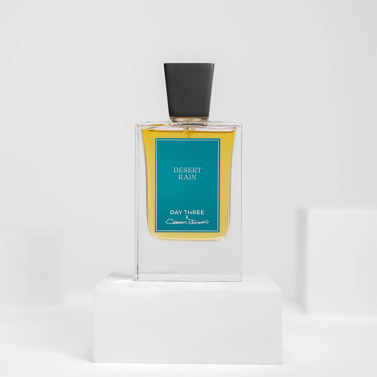 Day Three Desert Rain eau de parfum in a tall clear rectangular bottle with golden liquid, a wide angular matte-black cap and teal-blue rectangular label, centered on a white pedestal.