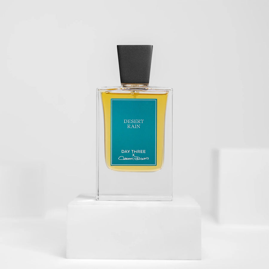 Day Three Desert Rain eau de parfum in a tall clear rectangular bottle with golden liquid, a wide angular matte-black cap and teal-blue rectangular label, centered on a white pedestal.