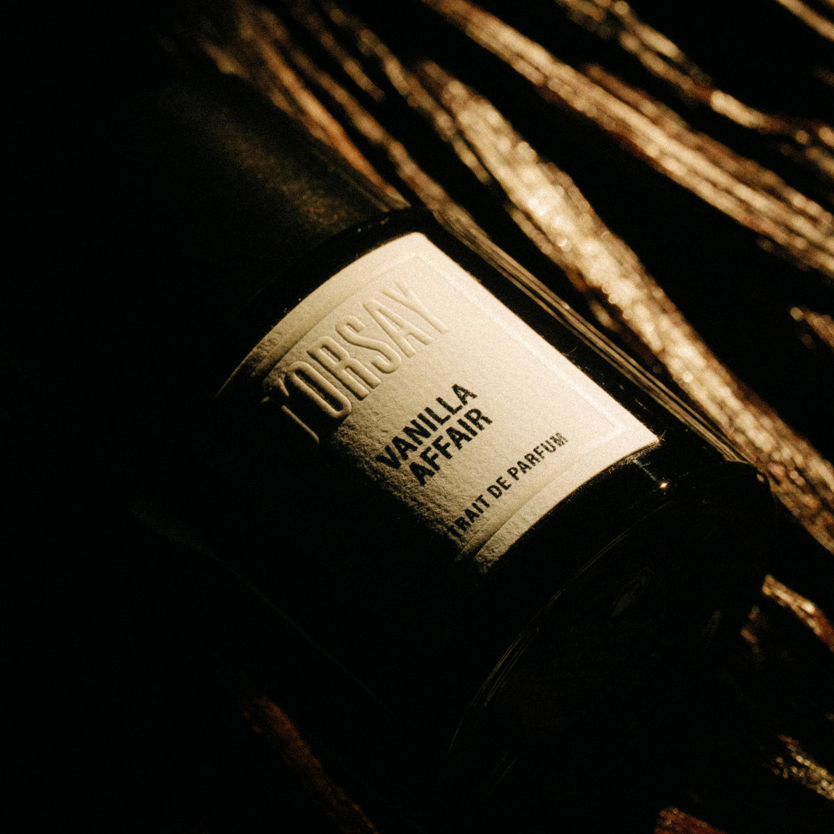 A close-up of a dark bottle labeled 'DORSAY VANILLA AFFAIR' with a cream label, resting on a dark wooden surface with dramatic warm lighting.