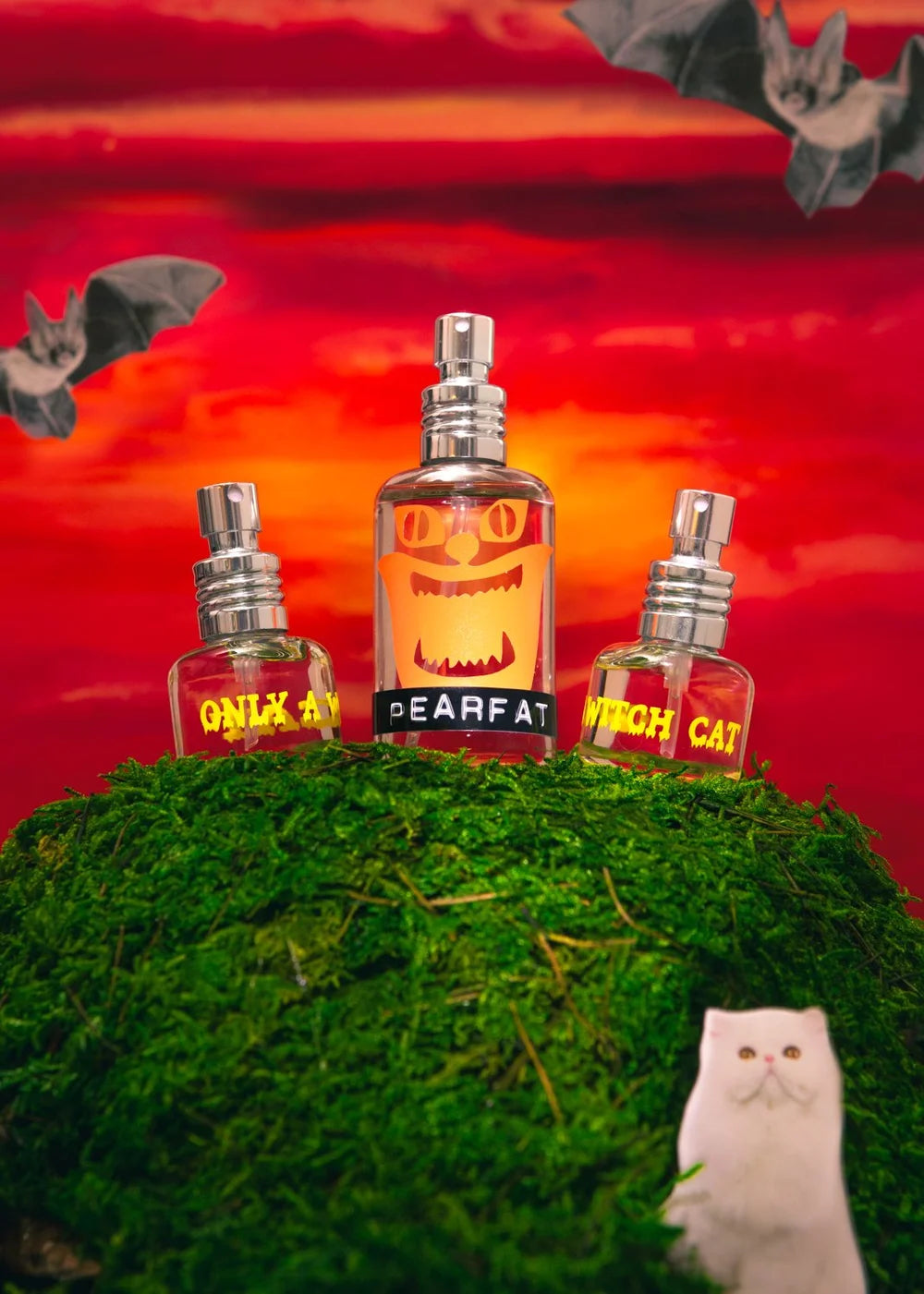 Three Pearfat Only a Witch Cat perfume bottles with exposed silver sprayers rising behind a mossy mound, with a white cat cutout, flying bats and a vivid red-orange painted sunset.
