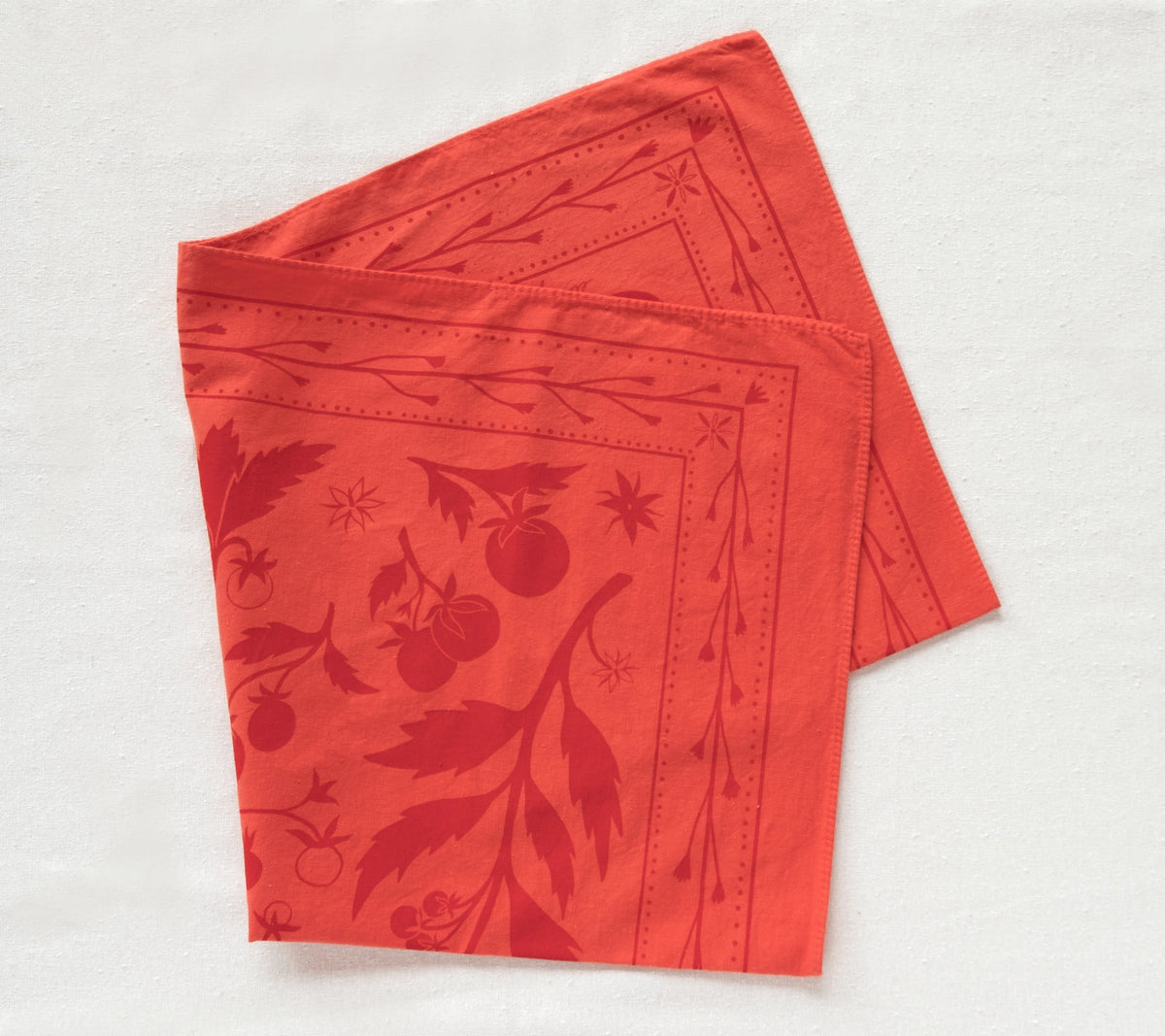 Two coral-red Elana Gabrielle Tomato Bandanas loosely folded on a pale linen surface, showing darker red tomato vines, fruit and border details.