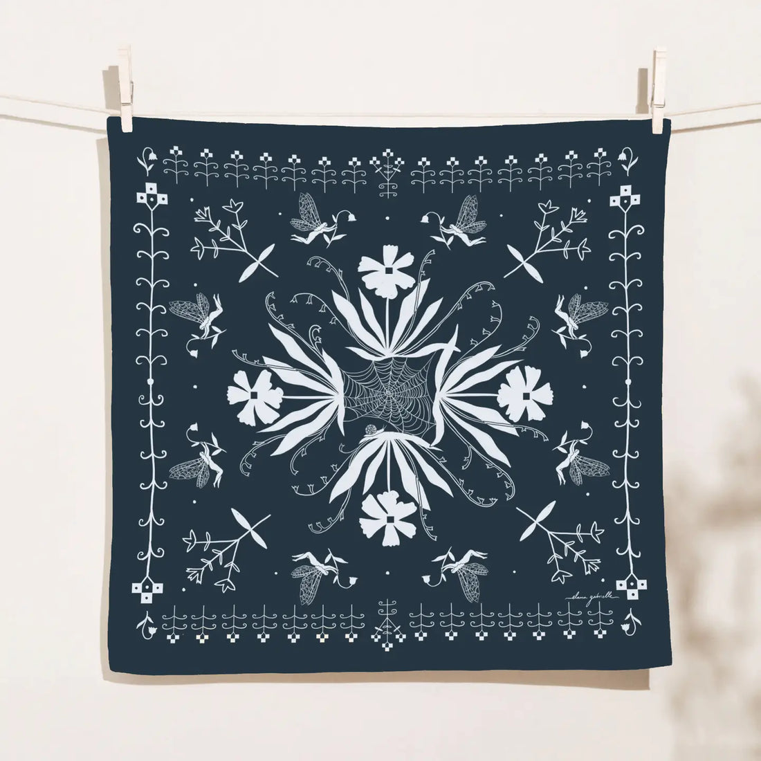 Dark navy square Hedgerow bandana clipped to a clothesline, printed in white with a symmetrical garden design of flowers, leafy stems, tiny winged figures and a central spiderweb motif.