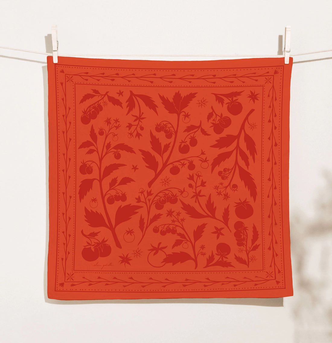 Coral-red Elana Gabrielle Tomato Bandana clipped to a clothesline, printed in darker red with sprawling tomato vines, leaves, blossoms and fruit inside a delicate botanical border.