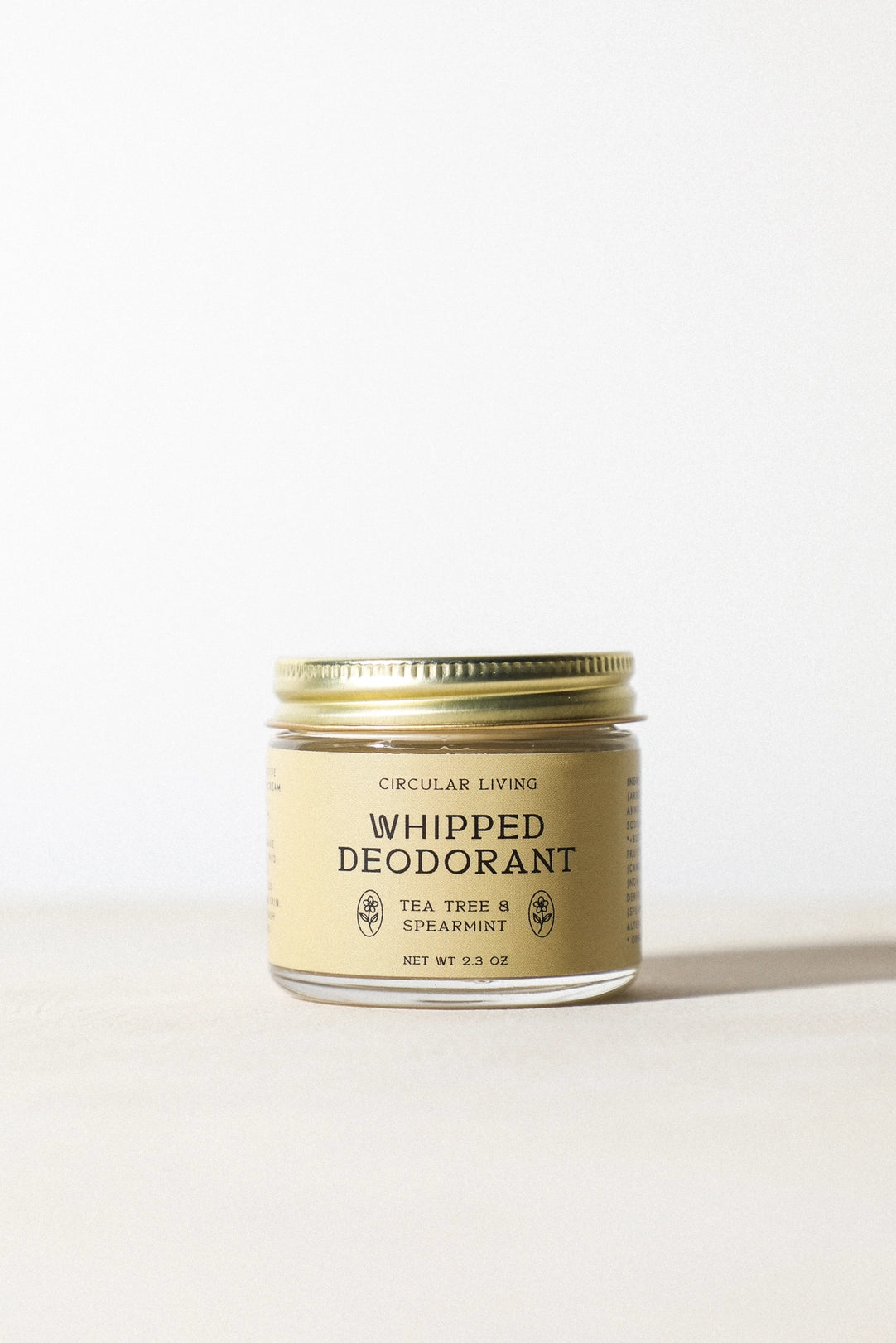 Circular Living Whipped Deodorant, Tea Tree & Spearmint in a small clear glass jar with pale cream label and gold metal screw lid, centered in warm sunlight on a light surface.