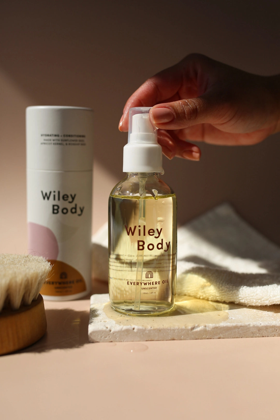 Wiley Body Everywhere Oil in a clear pump bottle filled with pale golden oil, held by a hand beside its tall white-and-blush carton, a dry body brush and folded towel in warm sunlight.